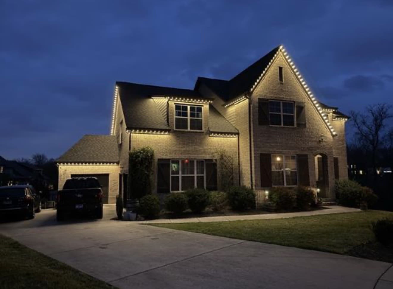 Two-story brick home with warm-white permanent lights tracing multiple steep gables and eaves.