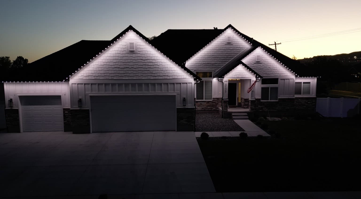 White board-and-batten home with stone accents and bright white permanent lighting tracing multiple gables above a three-car garage.