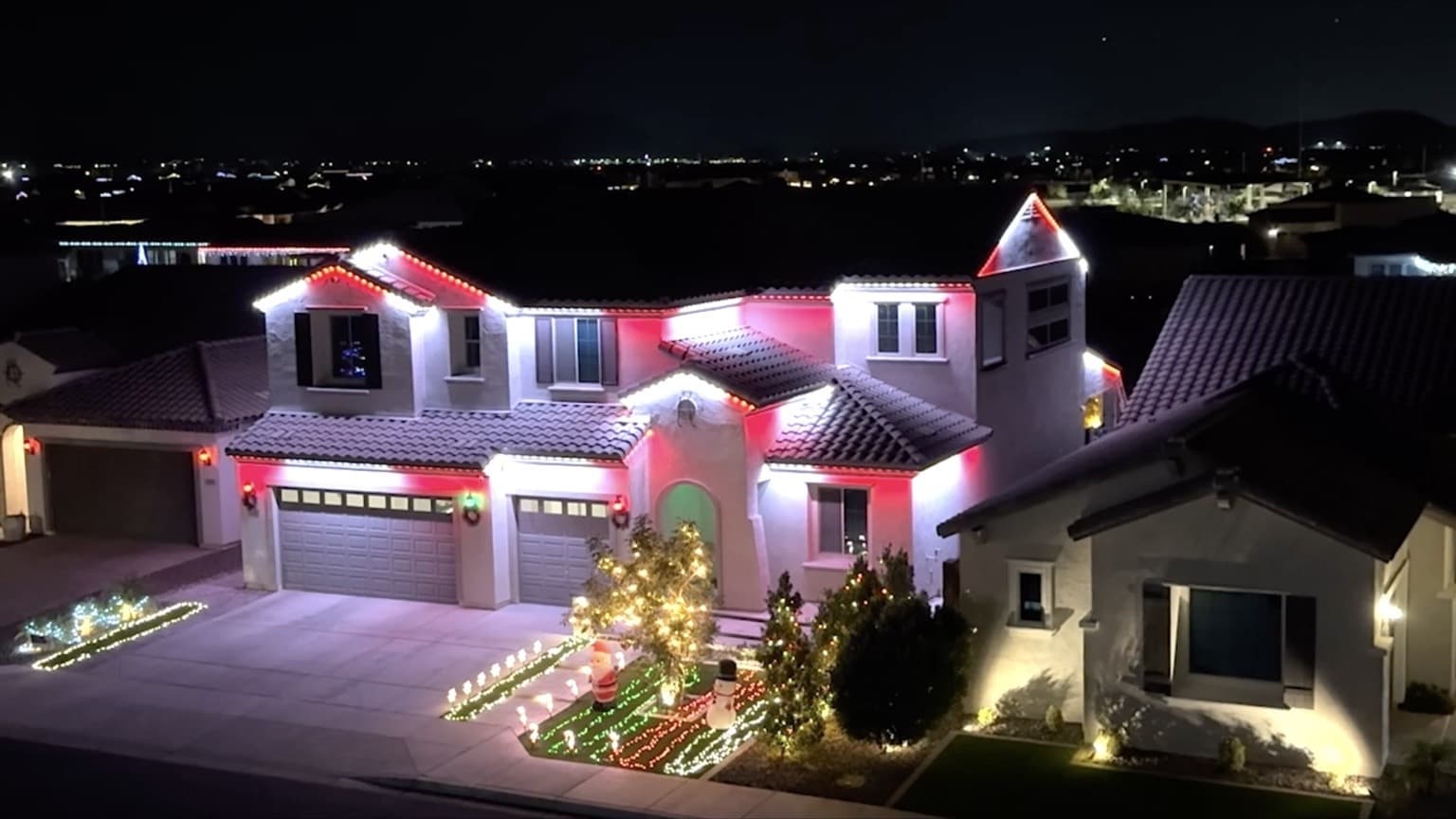 Large two-story stucco home with tile roof and red-and-white permanent lighting across multiple roof levels and garage bays.