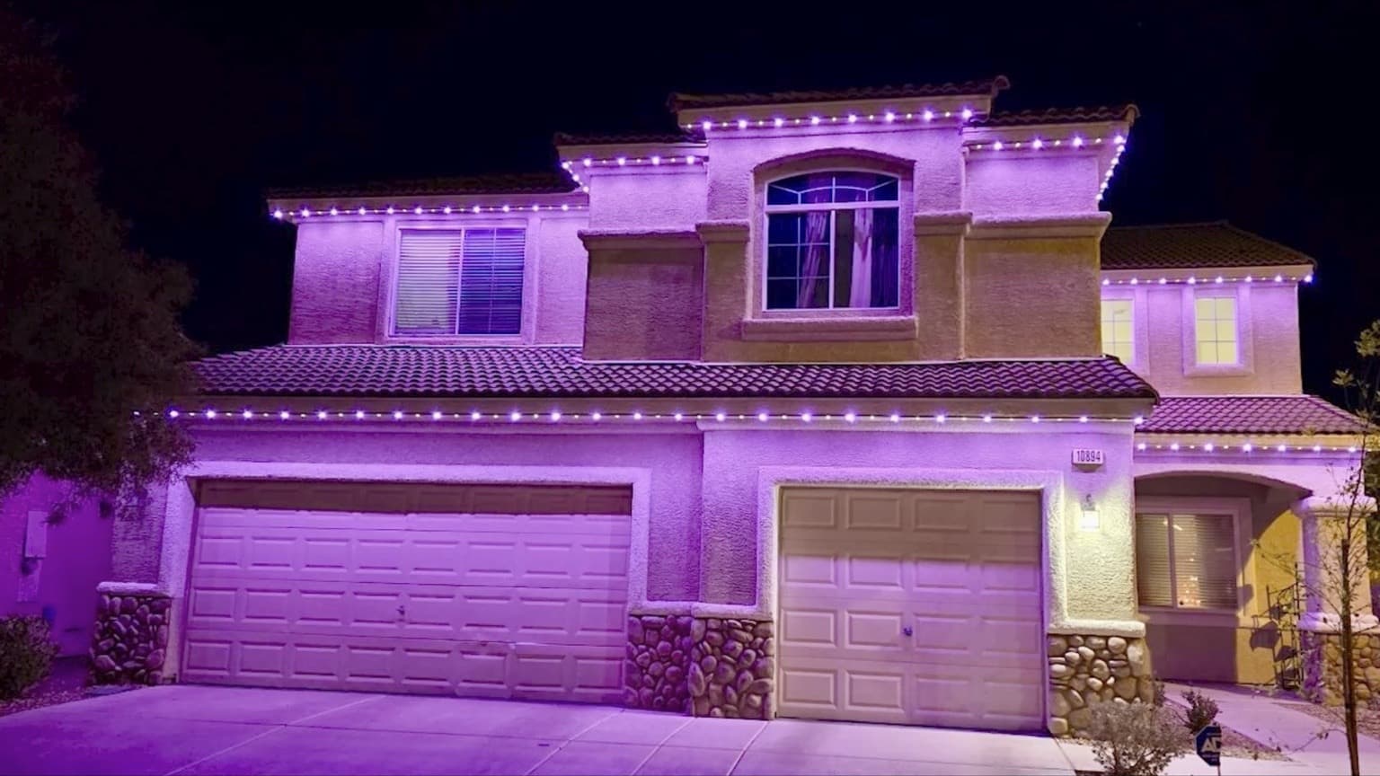 Two-story stucco home with tile roof and purple-and-white permanent lights tracing stepped rooflines above the garage and entry.