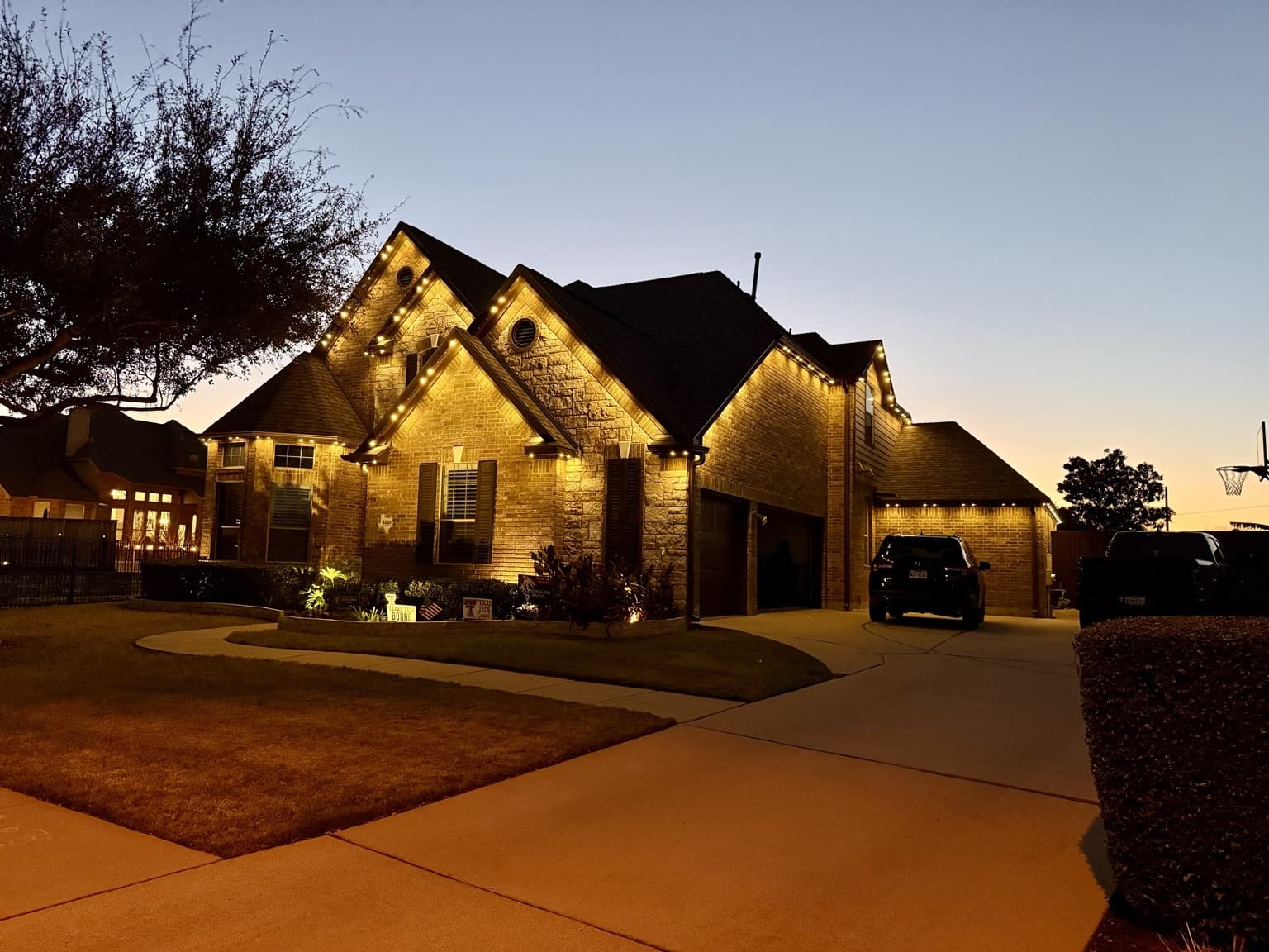 Brick-and-stone home with warm-white permanent lighting outlining layered gables and warm landscape accents near the entry.