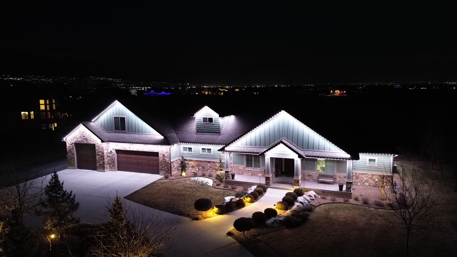 Elevated view of a stone-and-siding home with bright white permanent roofline lighting and warm landscape accents along the front walk.